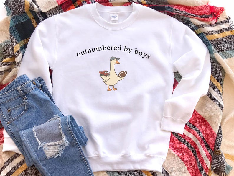 May include: White sweatshirt with the text "outnumbered by boys" above a cartoon goose wearing a hat and holding a football and a toy car. The sweatshirt is paired with blue jeans on a plaid blanket.