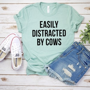 May include: A light blue t-shirt with the text "EASILY DISTRACTED BY COWS" in black lettering. The shirt is laid out on a white wooden surface with a pair of white canvas shoes and a pair of light blue denim shorts.