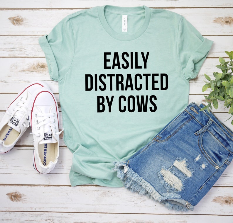May include: A light blue t-shirt with the text "EASILY DISTRACTED BY COWS" in black lettering. The shirt is laid out on a white wooden surface with a pair of white canvas shoes and a pair of light blue denim shorts.