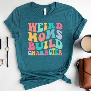 Pode incluir: Camiseta azul turquesa com o texto "Weird Moms Build Character" em um arco-&iacute;ris de cores. O texto est&aacute; em um estilo de fonte retr&ocirc;.