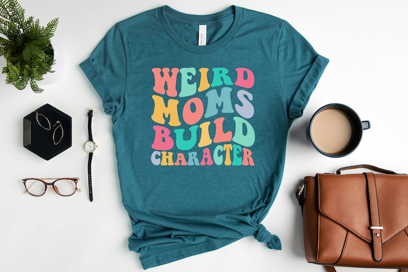 Pode incluir: Camiseta azul turquesa com o texto "Weird Moms Build Character" em um arco-&iacute;ris de cores. O texto est&aacute; em um estilo de fonte retr&ocirc;.