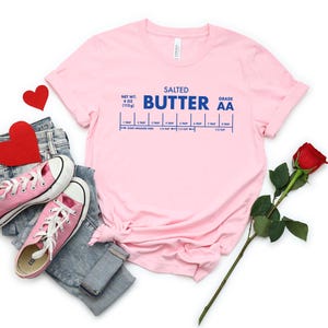 May include: Pink t-shirt with a blue graphic that reads "Salted Butter Grade AA" and a measurement guide for tablespoons and cups. The shirt is laid out on a white surface with a pair of pink sneakers, blue jeans, and a red rose.