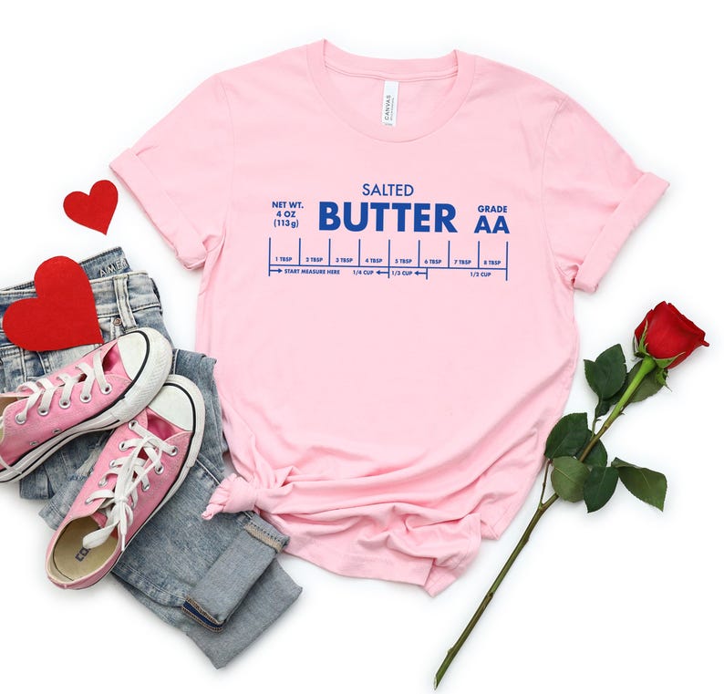 May include: Pink t-shirt with a blue graphic that reads "Salted Butter Grade AA" and a measurement guide for tablespoons and cups. The shirt is laid out on a white surface with a pair of pink sneakers, blue jeans, and a red rose.