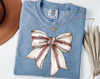May include: A light blue t-shirt with a white bow with red stitching, resembling a baseball. The shirt has a tag that reads "COMFORT COLORS" and "S - P - CH".