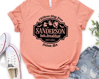 Puede incluir: Una camiseta de color melocotón con un gráfico negro de un letrero que dice "Sanderson Bed & Breakfast - Est. 1693 - Salem Ma". El letrero presenta una silueta negra de tres brujas volando en escobas.