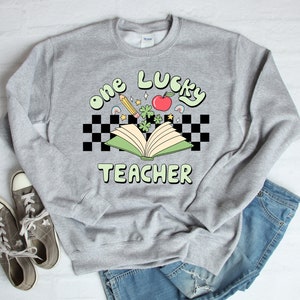 May include: A gray sweatshirt with the text "one lucky teacher" in green and white letters. The text is surrounded by a checkered pattern and a green open book with a red apple, shamrocks, and a pencil.