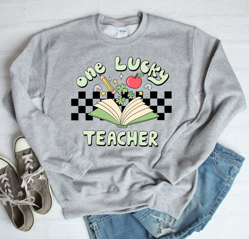 May include: A gray sweatshirt with the text "one lucky teacher" in green and white letters. The text is surrounded by a checkered pattern and a green open book with a red apple, shamrocks, and a pencil.