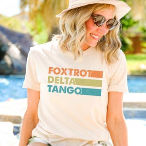 以下が含まれることがあります： カラフルなストライプでヴィンテージスタイルの「FOXTROT DELTA TANGO」という文字がプリントされた薄いベージュのTシャツを着た女性です。