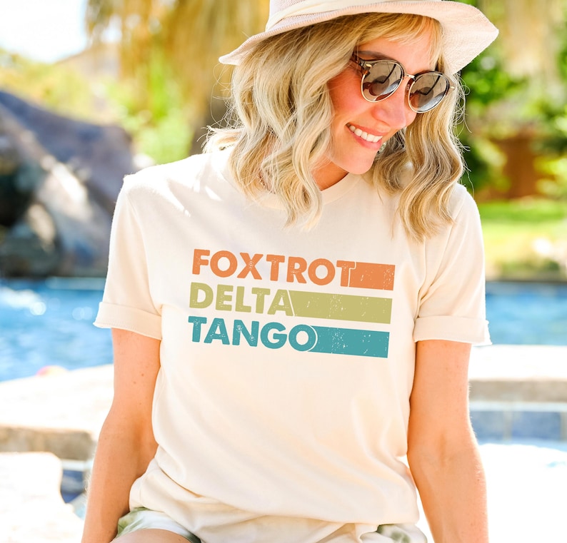 以下が含まれることがあります： カラフルなストライプでヴィンテージスタイルの「FOXTROT DELTA TANGO」という文字がプリントされた薄いベージュのTシャツを着た女性です。