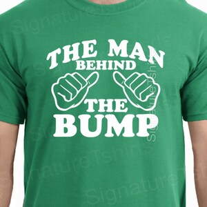 May include: Green t-shirt with white text that reads "The Man Behind The Bump" with two white thumbs up symbols.
