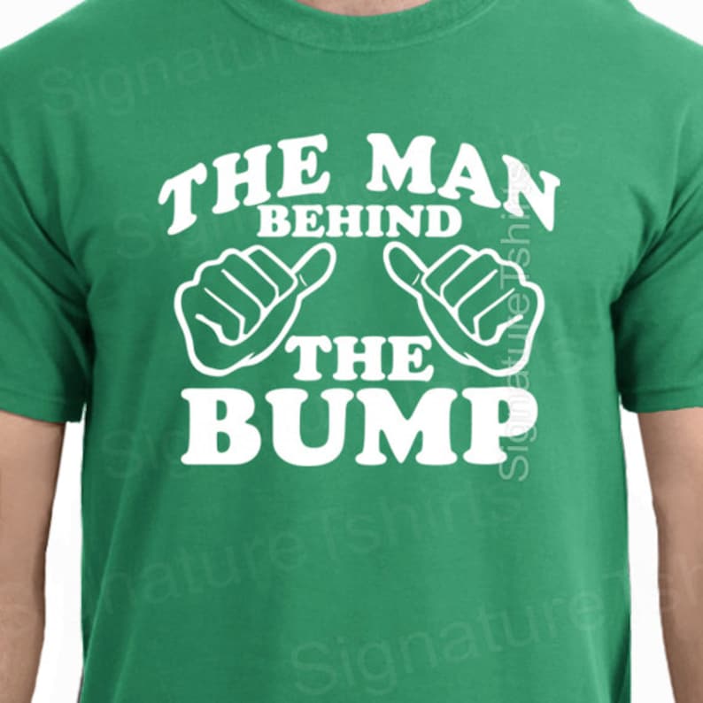 May include: Green t-shirt with white text that reads "The Man Behind The Bump" with two white thumbs up symbols.