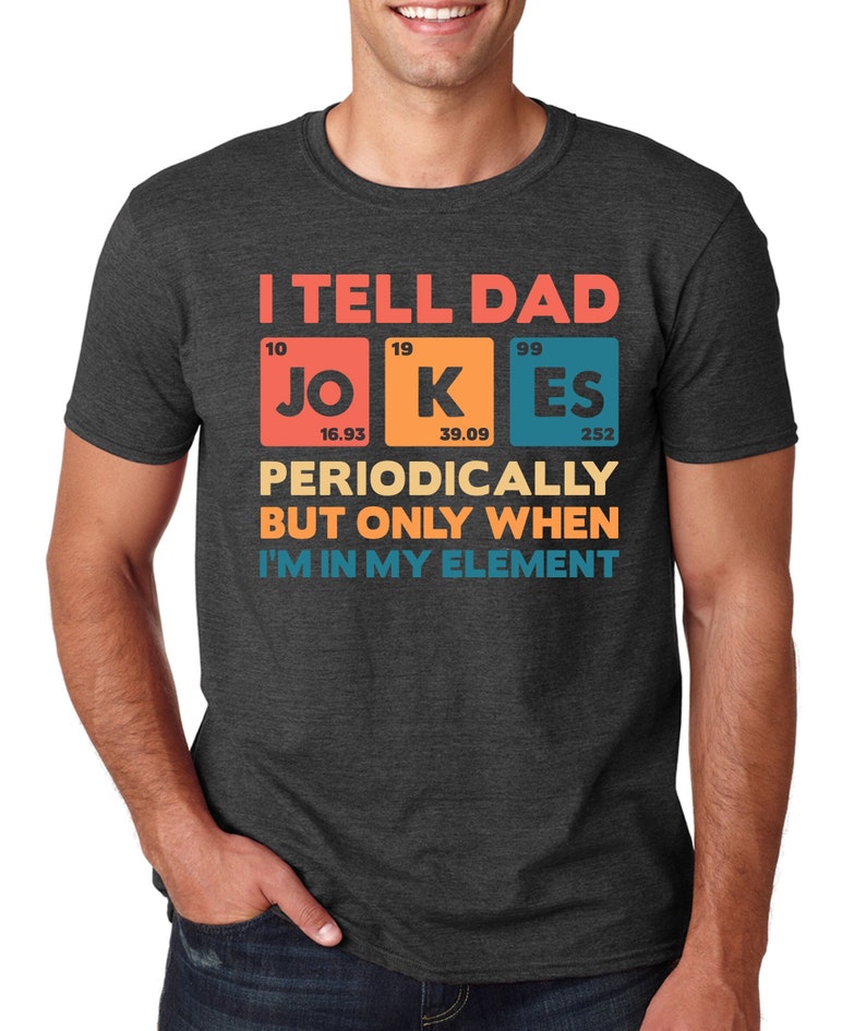 Puede incluir: Camiseta gris oscuro con el texto "I TELL DAD JOKES PERIODICALLY BUT ONLY WHEN I'M IN MY ELEMENT" en naranja, azul y amarillo. La palabra "JOKES" se muestra como elementos de la tabla peri&oacute;dica.