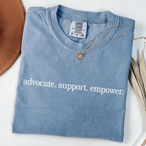 Op de afbeelding: Een blauw Comfort Colors T-shirt met de woorden "advocate. support. empower." in het wit. Een gouden ketting met een hanger ligt op het shirt. Een bruine hoed en gedroogde bladeren zijn op de achtergrond.