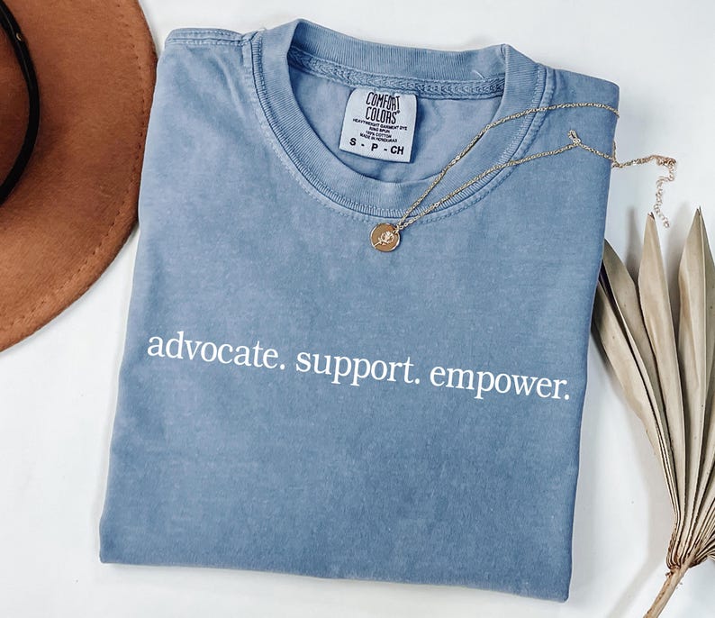 Op de afbeelding: Een blauw Comfort Colors T-shirt met de woorden "advocate. support. empower." in het wit. Een gouden ketting met een hanger ligt op het shirt. Een bruine hoed en gedroogde bladeren zijn op de achtergrond.