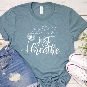 Op de afbeelding: Een tealblauw t-shirt met een witte paardenbloem en de tekst "just breathe" erop gedrukt.
