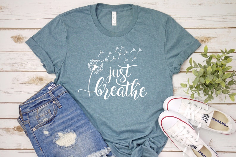 Op de afbeelding: Een tealblauw t-shirt met een witte paardenbloem en de tekst "just breathe" erop gedrukt.