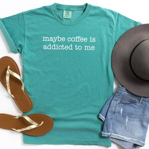 May include: Teal t-shirt with the phrase "maybe coffee is addicted to me" in white lettering. The shirt is paired with brown sandals with gold straps, denim shorts, and a gray hat. Casual wear.