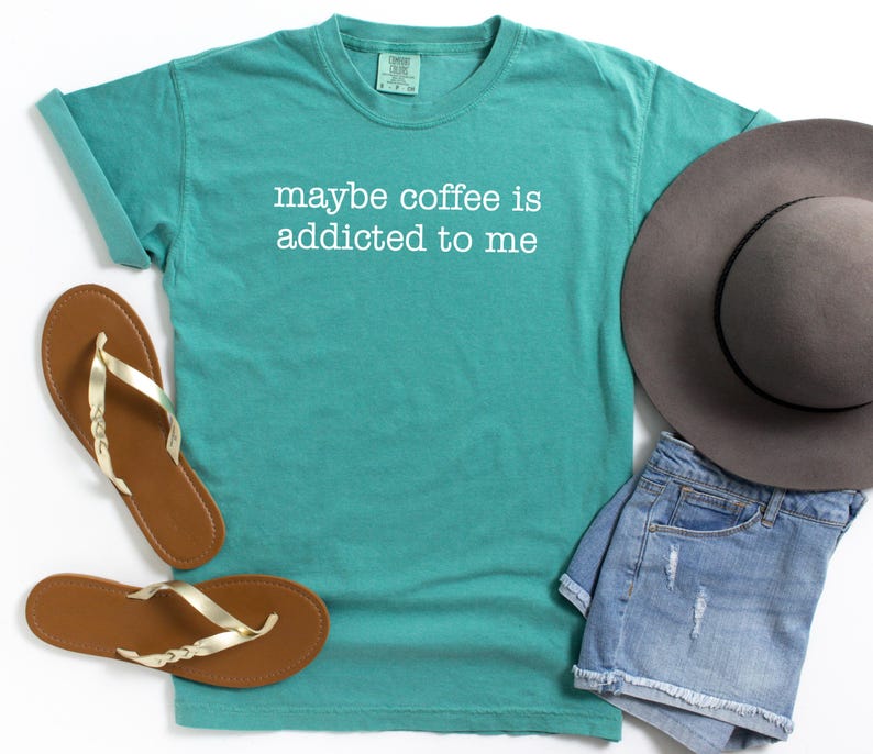 May include: Teal t-shirt with the phrase "maybe coffee is addicted to me" in white lettering. The shirt is paired with brown sandals with gold straps, denim shorts, and a gray hat. Casual wear.