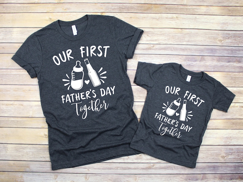 May include: Two dark gray t-shirts with white text and graphics. The larger shirt says "Our First Father's Day Together" with a baby bottle and a beer bottle graphic. The smaller shirt says "Our First Father's Day Together" with a baby bottle and a beer bottle graphic.
