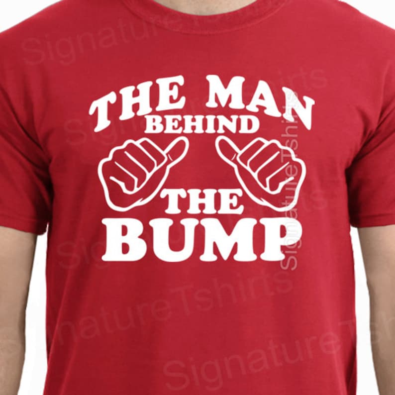 May include: Red t-shirt with white text that reads "The Man Behind The Bump" with two white thumbs up symbols.