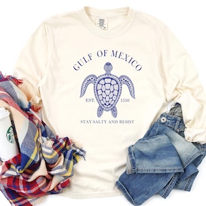 Op de afbeelding: Een cr&egrave;mekleurig longsleeve t-shirt met een blauwe afbeelding van een zeeschildpad en de tekst "Gulf of Mexico Est. 1550 Stay Salty and Resist".