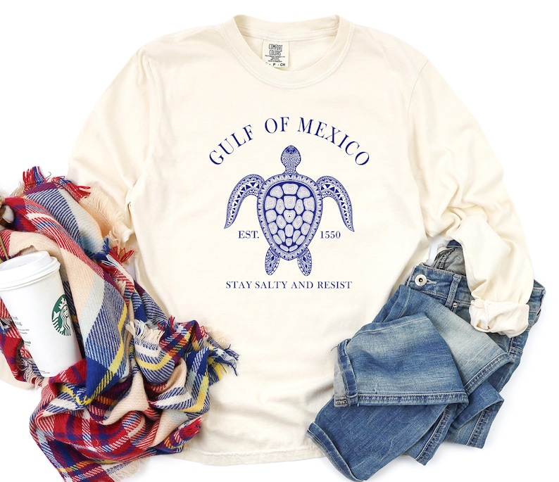 Op de afbeelding: Een cr&egrave;mekleurig longsleeve t-shirt met een blauwe afbeelding van een zeeschildpad en de tekst "Gulf of Mexico Est. 1550 Stay Salty and Resist".