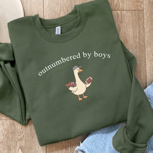 May include: An olive green sweatshirt with the phrase "outnumbered by boys" in white script. A cartoon goose wearing a baseball cap and holding a football and toy car is printed below the text. The sweatshirt is folded on a wooden surface.