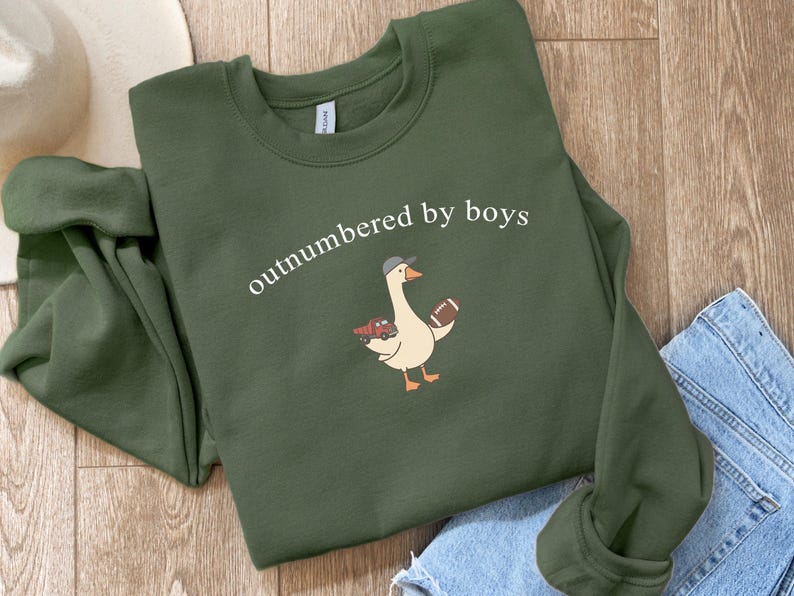 May include: An olive green sweatshirt with the phrase "outnumbered by boys" in white script. A cartoon goose wearing a baseball cap and holding a football and toy car is printed below the text. The sweatshirt is folded on a wooden surface.