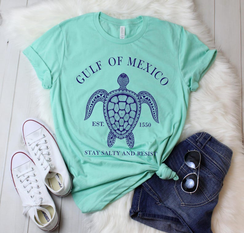 Puede incluir: Una camiseta verde claro con un dise&ntilde;o de tortuga azul marino y el texto "GULF OF MEXICO EST. 1550 STAY SALTY AND RESIST." Zapatillas blancas y pantalones cortos de mezclilla est&aacute;n al lado de la camisa.