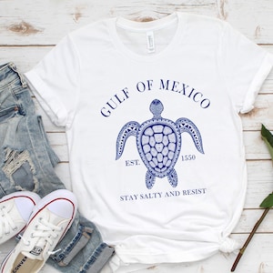 Puede incluir: Camiseta blanca con un dise&ntilde;o de tortuga azul y el texto "GULF OF MEXICO EST. 1550 STAY SALTY AND RESIST." La camiseta se combina con vaqueros de mezclilla de lavado claro y zapatillas blancas. Una rosa roja est&aacute; al lado.