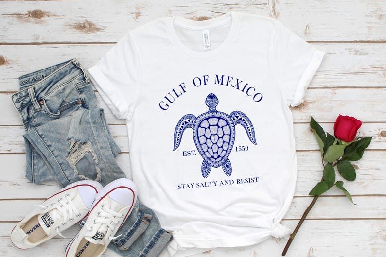 Puede incluir: Camiseta blanca con un dise&ntilde;o de tortuga azul y el texto "GULF OF MEXICO EST. 1550 STAY SALTY AND RESIST." La camiseta se combina con vaqueros de mezclilla de lavado claro y zapatillas blancas. Una rosa roja est&aacute; al lado.
