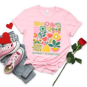 Op de afbeelding: Roze T-shirt met een afbeelding van kleurrijke bloemen, vlinders en bladeren. De tekst "COMMUNITY IS RESISTANCE" is onder de afbeelding gedrukt.
