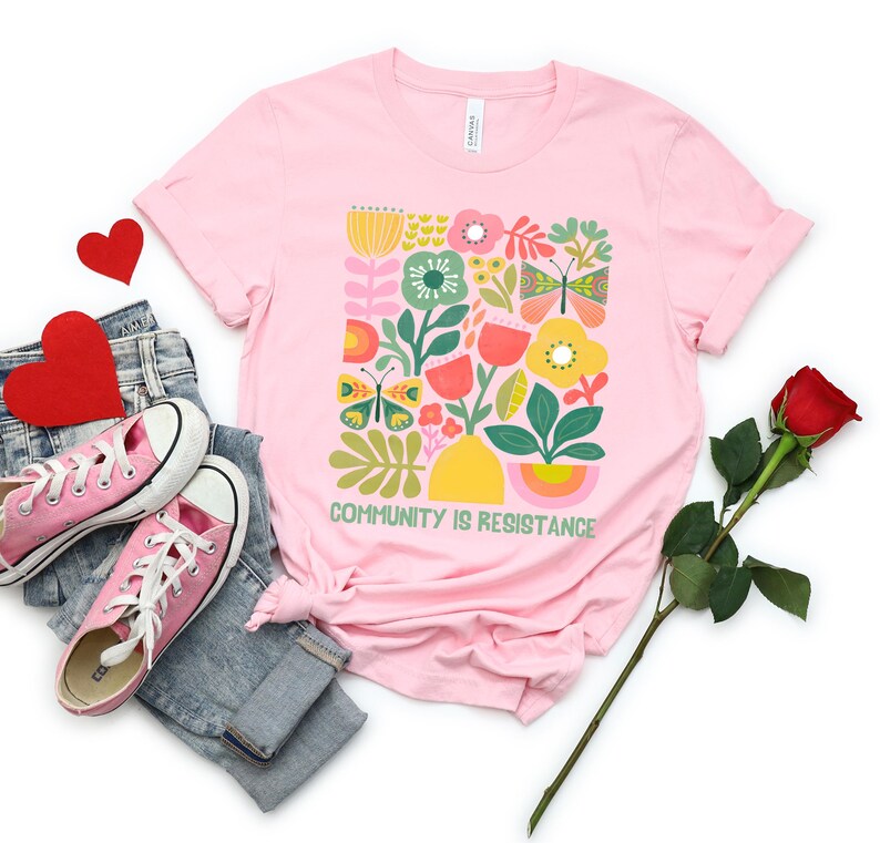 Op de afbeelding: Roze T-shirt met een afbeelding van kleurrijke bloemen, vlinders en bladeren. De tekst "COMMUNITY IS RESISTANCE" is onder de afbeelding gedrukt.