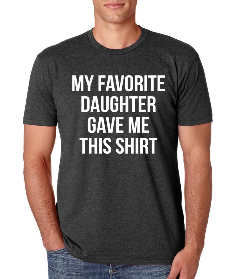 K&ouml;nnte beinhalten: Dunkelgraues T-Shirt mit dem wei&szlig;en Text "MY FAVORITE DAUGHTER GAVE ME THIS SHIRT". Das Shirt hat einen Rundhalsausschnitt und scheint aus einem weichen, bequemen Material zu sein.