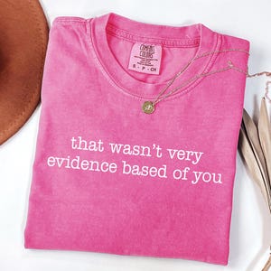 May include: A bright pink t-shirt with the phrase "that wasn't very evidence based of you" in white lettering. A gold necklace with a pendant rests on the shirt. The shirt is folded on a white surface.