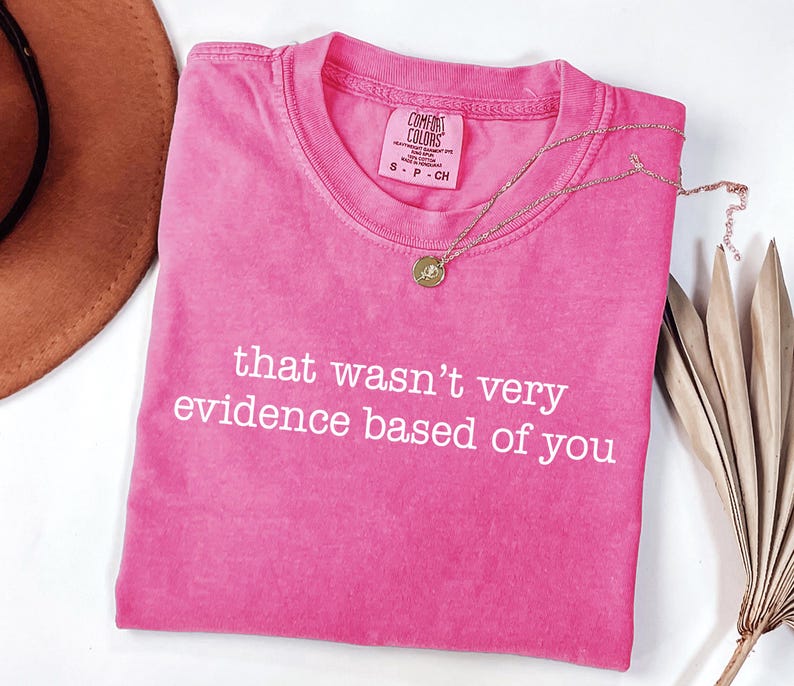 May include: A bright pink t-shirt with the phrase "that wasn't very evidence based of you" in white lettering. A gold necklace with a pendant rests on the shirt. The shirt is folded on a white surface.