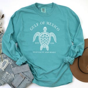 Op de afbeelding: Teal langarm T-shirt met een witte afbeelding van een zeeschildpad en de tekst "Gulf of Mexico Est. 1550 Stay Salty and Resist".