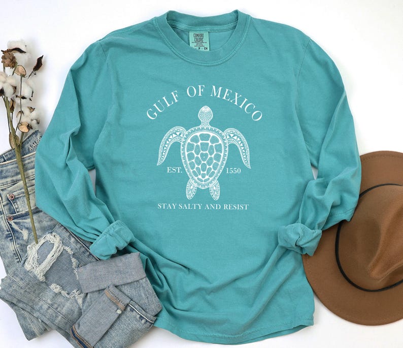 Op de afbeelding: Teal langarm T-shirt met een witte afbeelding van een zeeschildpad en de tekst "Gulf of Mexico Est. 1550 Stay Salty and Resist".