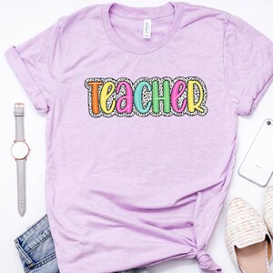 Puede incluir: Una camiseta lavanda con la palabra "Teacher" en letras de colores con un patr&oacute;n de lunares blancos.