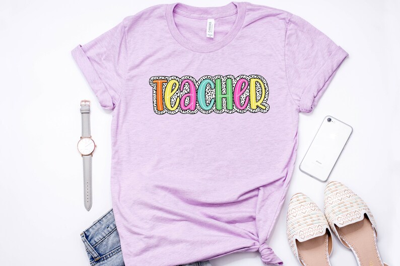 Puede incluir: Una camiseta lavanda con la palabra "Teacher" en letras de colores con un patr&oacute;n de lunares blancos.