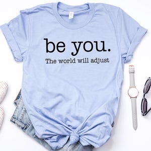 Puede incluir: Una camiseta azul claro con el texto "be you. The world will adjust" en negro. La camiseta se combina con un par de zapatos blancos, pantalones cortos de mezclilla, un reloj y gafas de sol. Los art&iacute;culos est&aacute;n colocados sobre una superficie blanca.