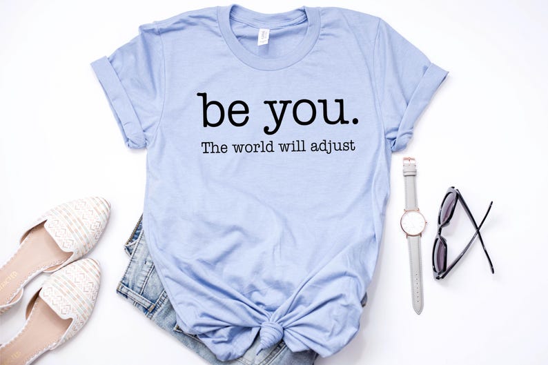 Puede incluir: Una camiseta azul claro con el texto "be you. The world will adjust" en negro. La camiseta se combina con un par de zapatos blancos, pantalones cortos de mezclilla, un reloj y gafas de sol. Los art&iacute;culos est&aacute;n colocados sobre una superficie blanca.