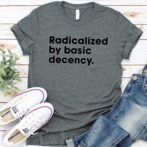 May include: A gray t-shirt with the text "Radicalized by basic decency." is paired with blue jeans and white sneakers. The sneakers have red accents and are placed near a small green plant on a wooden surface.