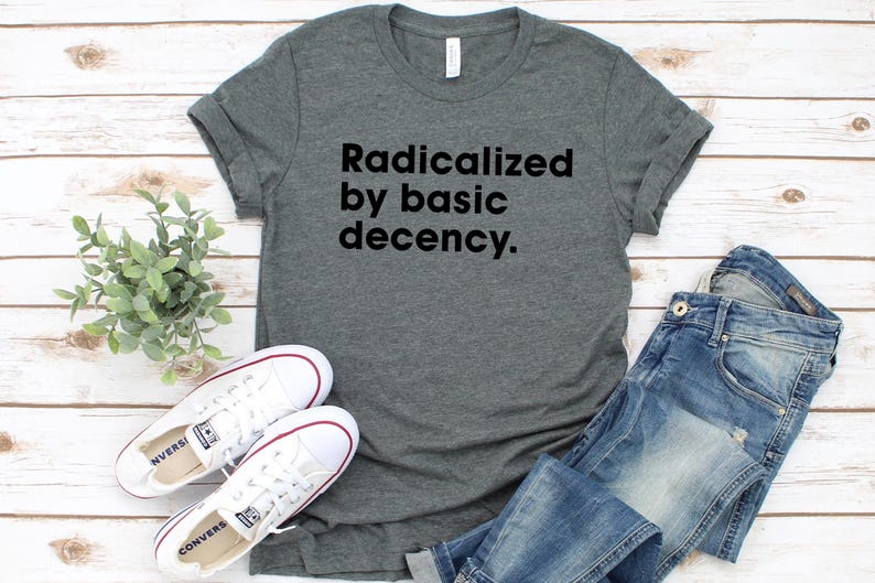 May include: A gray t-shirt with the text "Radicalized by basic decency." is paired with blue jeans and white sneakers. The sneakers have red accents and are placed near a small green plant on a wooden surface.