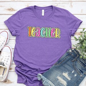 Puede incluir: Camiseta morada con la palabra "Teacher" en colores arco&iacute;ris con lunares blancos. La camiseta est&aacute; sobre un fondo de madera blanca con un par de zapatillas blancas y unos pantalones cortos de mezclilla azul.
