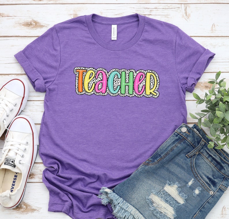 Puede incluir: Camiseta morada con la palabra "Teacher" en colores arco&iacute;ris con lunares blancos. La camiseta est&aacute; sobre un fondo de madera blanca con un par de zapatillas blancas y unos pantalones cortos de mezclilla azul.