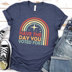 May include: Navy blue t-shirt with a retro rainbow graphic and the text "Have the day you voted for." The shirt is paired with blue jeans, black sneakers, and a watch. The shirt is made of a soft, comfortable material.