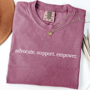 Op de afbeelding: Een mauve Comfort Colors t-shirt met de woorden "advocate. support. empower." in het wit. Een gouden ketting met een hanger en een bruine hoed zijn ook zichtbaar.