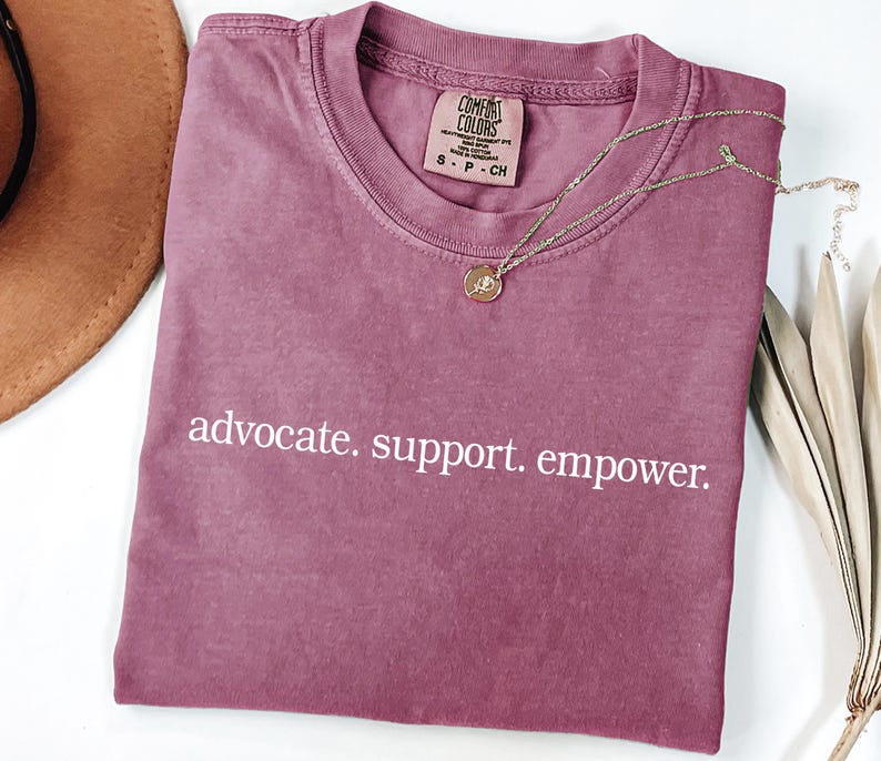 Op de afbeelding: Een mauve Comfort Colors t-shirt met de woorden "advocate. support. empower." in het wit. Een gouden ketting met een hanger en een bruine hoed zijn ook zichtbaar.