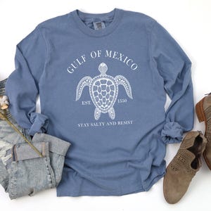 Op de afbeelding: Een blauw longsleeve t-shirt met een witte afbeelding van een zeeschildpad en de tekst "Gulf of Mexico Est. 1550 Stay Salty and Resist".